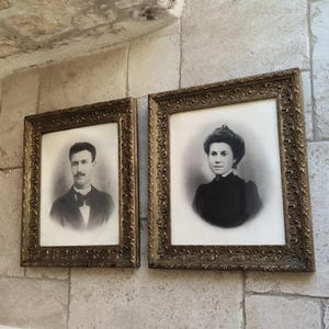 Peut inclure: Deux portraits encadrés sont accrochés à un mur de pierre. Les portraits sont en noir et blanc, représentant un homme et une femme. Les cadres sont ornés, avec une finition dorée. L'homme porte un costume et un nœud papillon. La femme porte une robe à col haut.