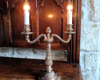 May include: A gold-toned, ornate, three-armed candelabra with two lit candles. The candelabra has a decorative base and is on a wooden surface.
