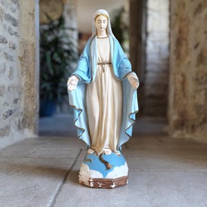 Może przedstawiać: Stojąca figura Najświętszej Maryi Panny w kremowej szacie i jasnoniebieskim welonie ze złotym wykończeniem. Postać ma wyciągnięte dłonie i stoi na niebieskiej podstawie w kształcie globu z białymi chmurami oraz wężem owiniętym u dołu figury.