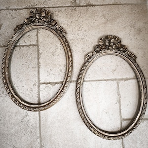 May include: Two antique oval picture frames with ornate gold-colored detailing. Each frame features a decorative bow at the top and a textured border. The frames are empty, ready for a photo or artwork.