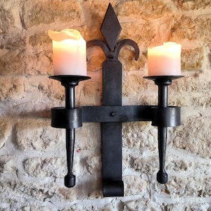 Peut inclure: Un bougeoir mural en métal noir avec deux bras, chacun tenant une bougie blanche. Le bougeoir a un motif décoratif de fleur de lys.