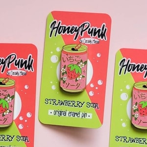以下が含まれることがあります： イチゴソーダのピンクの缶を特徴とするエナメルピンが3つ。缶には「いちごソーダ」と日本語で書かれています。各ピンは、「Honey Punk」と「STRAWBERRY SODA - original enamel pin.」の文字が書かれた緑とピンクのカードの上にあります。