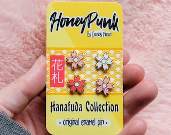 May include: A set of three enamel pins featuring pink and white cherry blossoms. The pins are on a yellow card with the text "HoneyPunk by Cassidy Mason" and "Hanafuda Collection - original enamel pin - ".