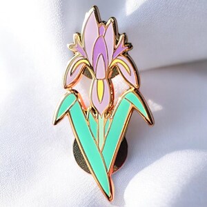 Peut inclure: Une épingle en émail dorée représentant une fleur d'iris stylisée de couleur violette et jaune avec des feuilles vertes.