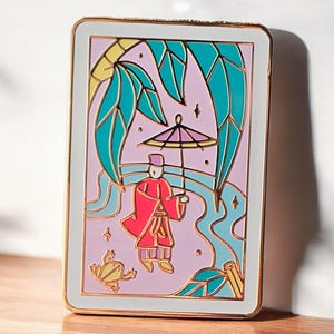 以下が含まれることがあります： 白い背景に金色のエナメルピン。赤いローブを着た人が傘を持って立っている漫画のイラストが描かれており、緑色のカエルとヤシの木のあるピンクとブルーの風景の中にいる。