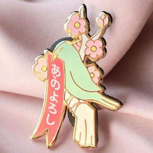 May include: A green bird with a red ribbon that says "Let's go!" in Japanese is perched on a branch with pink flowers. The bird and branch are gold-coloured.