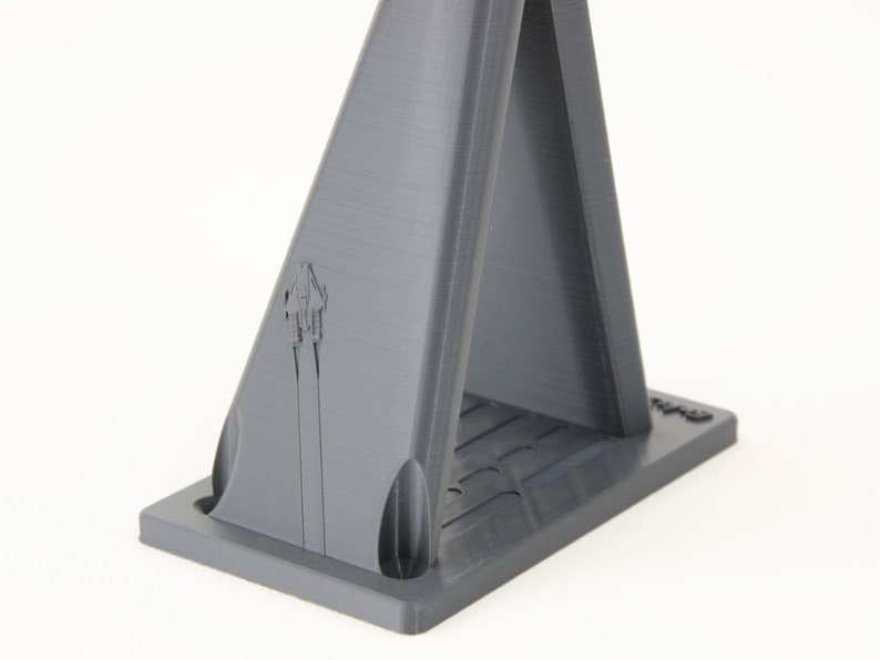 May include: A gray 3D printed stand with a triangular base and two vertical supports. The stand has a small logo on the front.