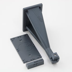 May include: A gray 3D printed wall mount with a rectangular base and a triangular shaped arm. The arm has a small square connector at the end. The base has a small hole in the center and a logo with the text "Battery 5.2cm".