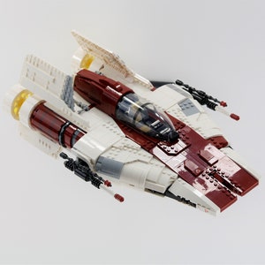 May include: A white and red Lego spaceship with a clear cockpit and black guns. The spaceship has a yellow engine and is on a white background.