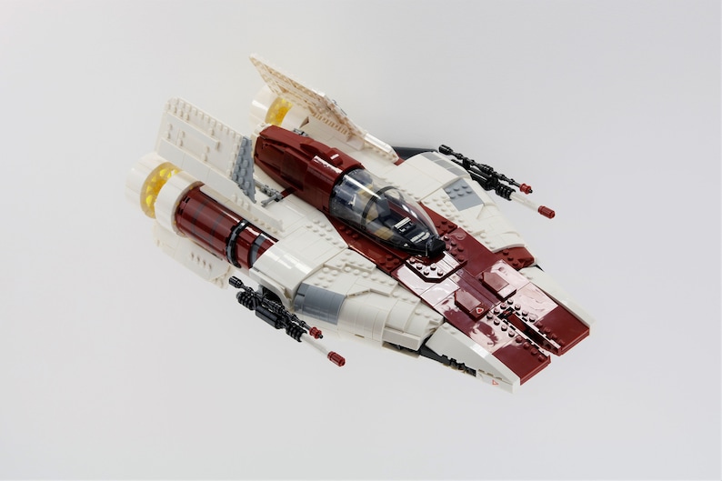 May include: A white and red Lego spaceship with a clear cockpit and black guns. The spaceship has a yellow engine and is on a white background.