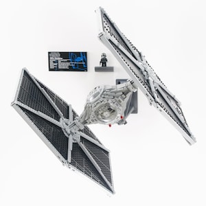 May include: A detailed model of a TIE Fighter from Star Wars, constructed from gray and black building blocks. The model includes the iconic wing panels and central cockpit. A small black figure and a black information plaque are also present.