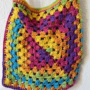 May include: A handmade, colorful crochet bag with a square pattern. The bag features a rainbow of colors, including yellow, orange, pink, purple, blue, and green. The strap and trim are also crocheted. The bag is a vibrant and unique accessory.