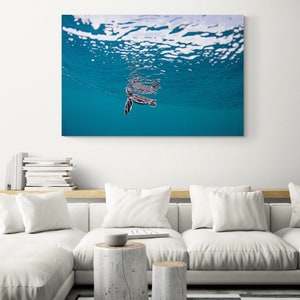 May include: A small sea turtle swims just below the surface of the water. The water is a bright blue and the turtle is dark grey with black markings. The surface of the water is rippled and reflects the sunlight.
