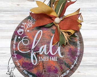 May include: A round wooden sign with the word "Fall" in white script, accented with a bow of orange, green, and yellow ribbons. The sign has a painted background with fall colors and the words "Sweet Fall".