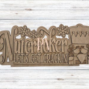 May include: Wooden sign with the words "Nutcracker" and "Let's Get Crackin'" in a layered design. The sign features snowflakes and a nutcracker soldier illustration. The sign is made of light brown wood.
