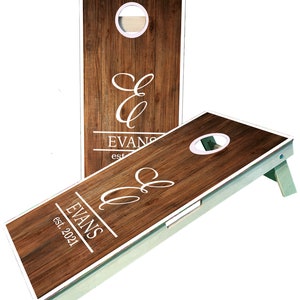 May include: A wooden cornhole set with a white frame and a brown woodgrain design. The set features a personalized monogram with the initials "E & E" and the last name "EVANS" with the text "est. 2021".