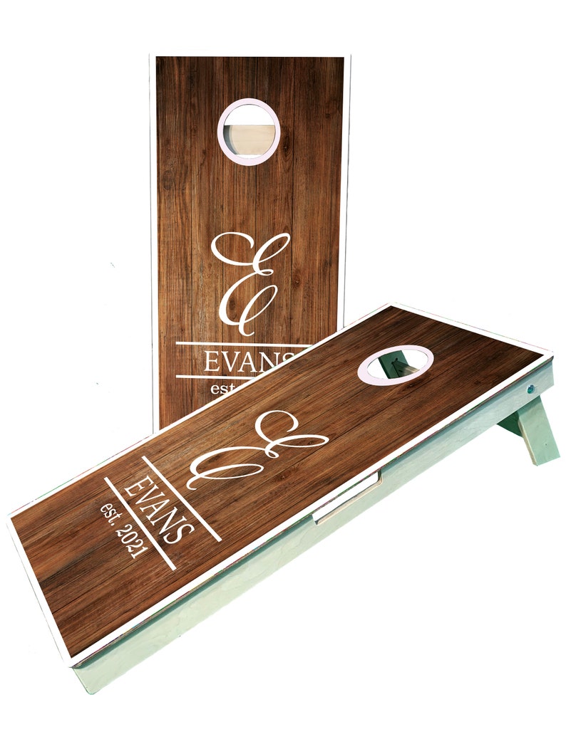 May include: A wooden cornhole set with a white frame and a brown woodgrain design. The set features a personalized monogram with the initials "E & E" and the last name "EVANS" with the text "est. 2021".