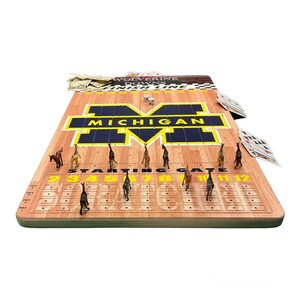 May include: A wooden game board with a Michigan Wolverines theme. The board features a yellow and blue 'M' logo and a starting gate with numbered spaces. There are miniature horses on the board, along with dice and playing cards.