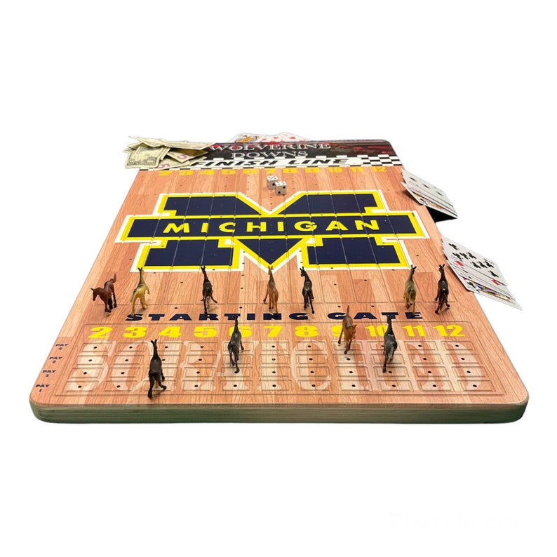 May include: A wooden game board with a Michigan Wolverines theme. The board features a yellow and blue 'M' logo and a starting gate with numbered spaces. There are miniature horses on the board, along with dice and playing cards.