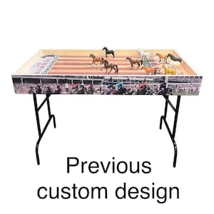 May include: A wooden horse racing game table with a black metal frame. The table top features a colorful racetrack design with a crowd of people watching the race. There are several plastic horses on the track.