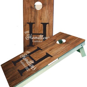 May include: A wooden cornhole set with a personalized design. The boards are brown with a wood grain texture and feature a large black letter "H" with the text "The Hamiltons Established 2022".