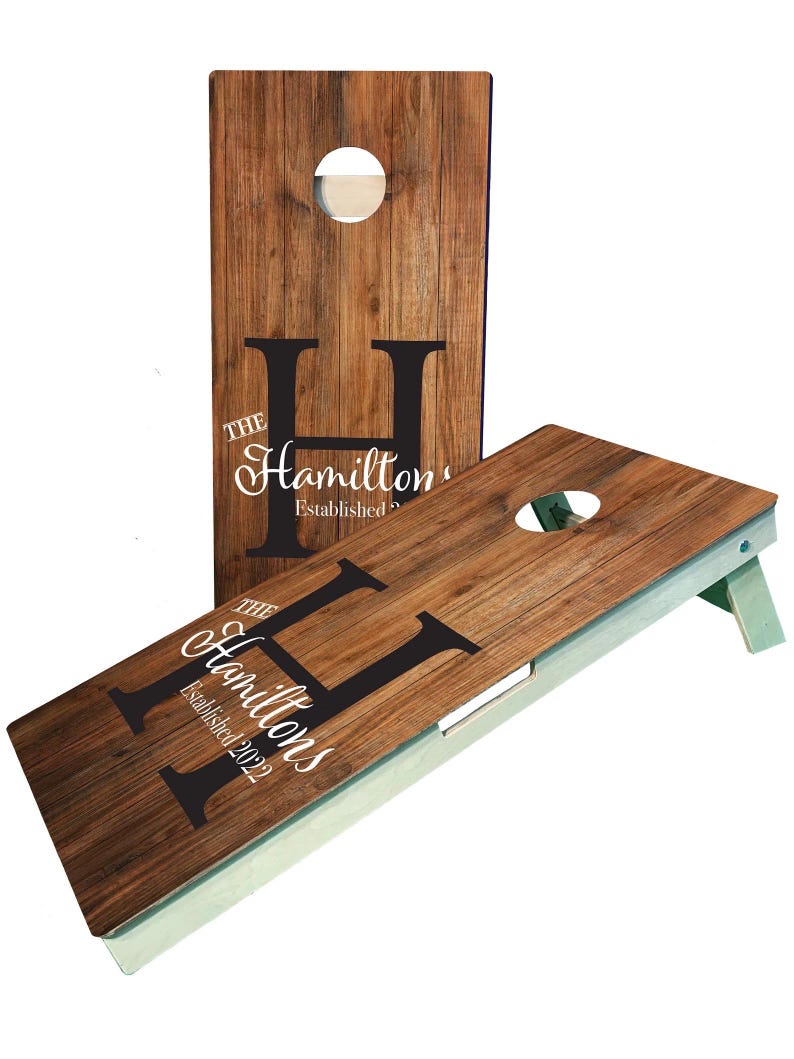 May include: A wooden cornhole set with a personalized design. The boards are brown with a wood grain texture and feature a large black letter "H" with the text "The Hamiltons Established 2022".