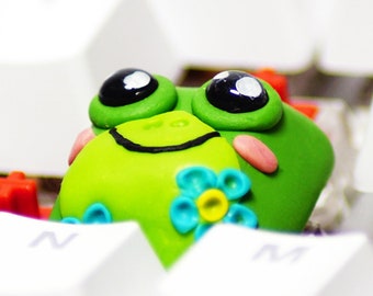 May include: A novelty green frog keycap with large black eyes and a smiling mouth rests on a white keyboard. The frog has pink cheeks and a blue flower design. The keycap adds a playful touch to any mechanical keyboard.