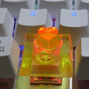 Peut inclure: Gros plan sur un clavier mécanique blanc avec des touches éclairées. Le clavier est équipé d'une touche personnalisée en forme de cœur orange. Les touches sont rétroéclairées en différentes couleurs, créant une esthétique vibrante.