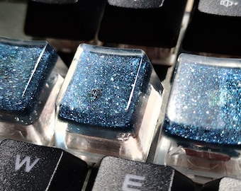 May include: Close-up of a mechanical keyboard with custom keycaps. The keycaps are a mix of black and clear, with some featuring a blue glitter design. The keyboard also has function keys and letter keys.