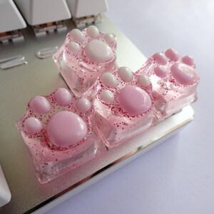 Four pink and white paw-shaped keycaps with glitter. The keycaps are made of clear plastic with pink and white paw prints inside. The keycaps are arranged in a row on a keyboard.