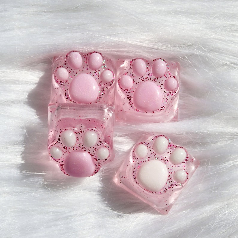 Four pink and white paw print keycaps with glitter. The keycaps are made of clear resin with pink and white glitter embedded in the resin. The paw prints are raised and have a three-dimensional appearance.