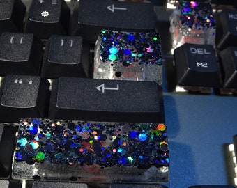 May include: Custom mechanical keyboard keycaps featuring a clear resin base embedded with chunky, multi-colored holographic glitter in shades of blue, green, and purple, contrasting against standard black keyboard keys.