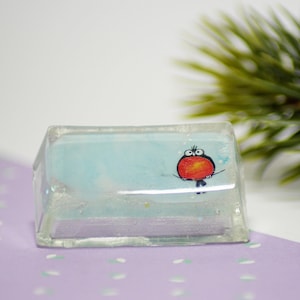 Puede incluir: Un pisapapeles rectangular de resina transparente con un interior azul claro. Un pájaro de dibujos animados con pecho rojo y detalles negros aparece en la parte delantera. El pisapapeles es un artículo decorativo.