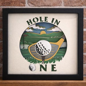 May include: Framed artwork with a golf theme. The design features a golf ball on a tee, a golf club, and the words "HOLE IN ONE." The background depicts a golf course scene with a flag, green grass, and a blue sky. The artwork is in a black frame.