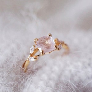 Op de afbeelding: Een gouden ring met een lichtroze vierkante edelsteen als middelpunt. De ring heeft delicate, bladachtige accenten met kleine, heldere edelstenen. De ring is geplaatst tegen een zachte, witte achtergrond.