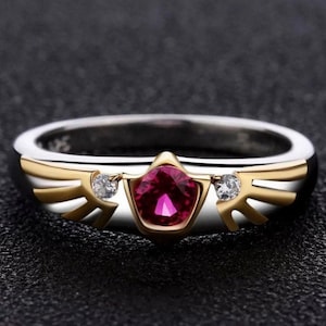May include: A two-tone ring with a gold and silver band. The ring features a central, faceted, deep pink gemstone, flanked by two smaller clear stones. Gold-colored wing-like accents are on either side of the central stone.