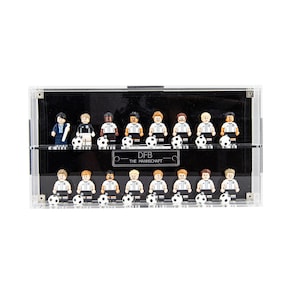 May include: A clear display case containing miniature football player figures. Each figure wears a white and black uniform and holds a football. The text "DFB THE MANNSCHAFT" is visible on the case.