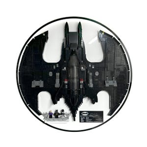 Op de afbeelding: Een zwart Batwing-model gemaakt van speelgoedblokken, gepresenteerd in een cirkelvormige lijst. Het model bevat een cockpit, vleugels en kleine figuren. Een zwarte informatiekaart is zichtbaar, met de tekst "Batwing (1989)".
