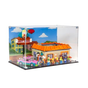 Peut inclure: Un ensemble Lego Krusty Burger présenté dans un boîtier en acrylique transparent. L'ensemble comprend une voiture rose, un bâtiment avec un toit orange et plusieurs figurines Lego. Le panneau indique "Krusty Burger".