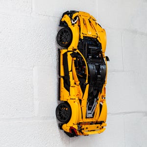 May include: A yellow Lego model of a McLaren Senna sports car, with black details, is mounted on a white wall.