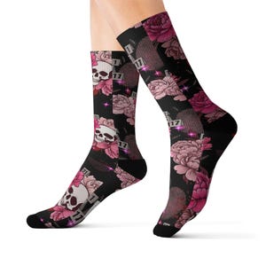 May include: A pair of black crew socks featuring a repeating pattern of white skulls, pink and dark pink peony flowers, and small rectangular tags with the number 17 printed on them, accented by small purple starbursts throughout the design.