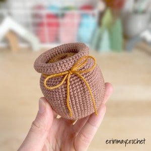 May include: A small crocheted pot with a brown yarn and a yellow ribbon tied around the top. The pot is being held in a hand.
