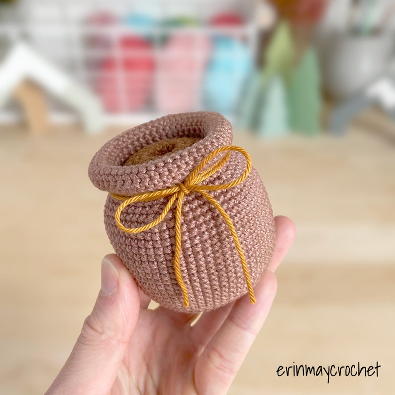 May include: A small crocheted pot with a brown yarn and a yellow ribbon tied around the top. The pot is being held in a hand.