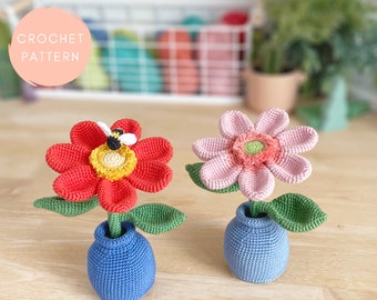 Puede incluir: Dos arreglos florales de crochet en macetas azules. Una flor es roja con un centro amarillo y una abeja en la parte superior. La otra flor es rosa con un centro coral.