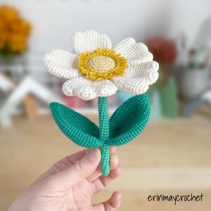 May include: A crocheted white flower with a yellow center and green leaves. The flower is held in a hand.