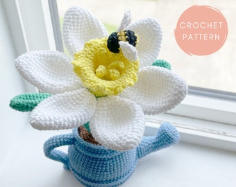 Può includere: Un narciso all'uncinetto con petali bianchi, centro giallo e una piccola ape. Il fiore è innaffiato da un annaffiatoio blu. Il testo "CROCHET PATTERN" è in un cerchio rosa. È visibile anche il nome dell'artista, @erinmaycrochet.