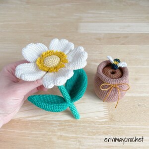 May include: A crocheted white flower with a yellow center and green leaves, next to a crocheted brown honey pot with a bee on top. The honey pot has a brown ribbon tied around it.