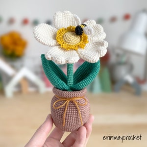 May include: A crocheted white flower with a yellow center and a bee on top, in a brown pot with a yellow ribbon.