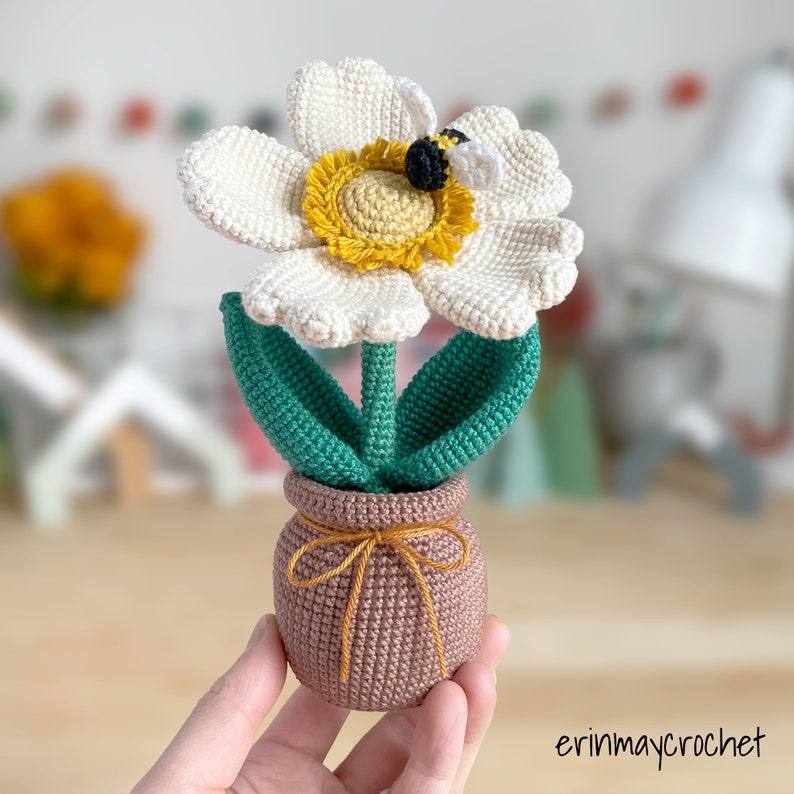 May include: A crocheted white flower with a yellow center and a bee on top, in a brown pot with a yellow ribbon.