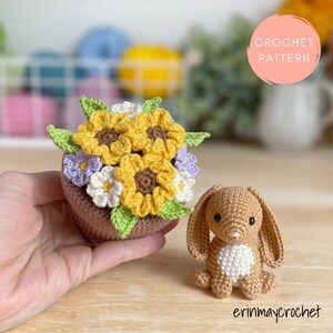 May include: A crocheted arrangement of yellow sunflowers, white daisies, and purple flowers in a brown pot. A small, tan crocheted bunny with long ears and a white belly sits beside the flowers. The image includes the text "CROCHET PATTERN" and "erinmaycrochet".